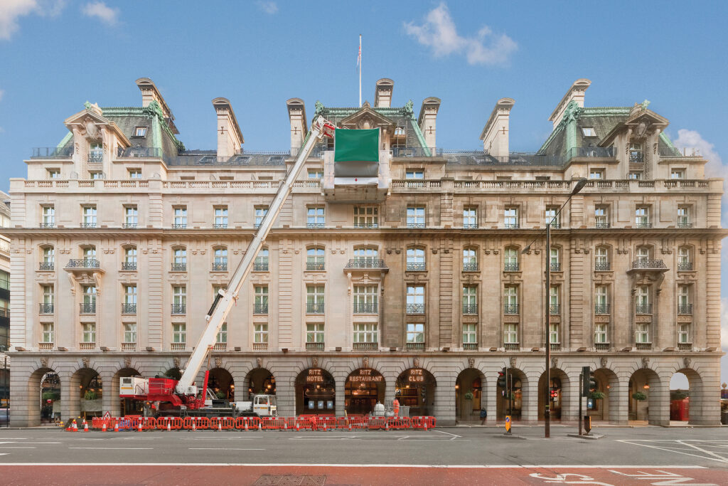Full frontage of The Ritz Hotel, London, featuring its Belle Époque Portland stone and Cornish grey granite façade, mansard roof with copper detailing, and arcaded entrance reading 'Ritz Hotel,' 'Ritz Restaurant,' and 'Ritz Club.' A red telescopic crane reaches up to the roofline, its shrouded platform positioned at the upper storey, with barriers and cones lining the pavement below. This equipment captures the Façade Gommage cleaning process underway, conducted by Styles Façade Solutions (formerly Thomann-Hanry®).