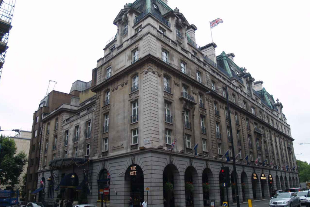 Wide corner view of The Ritz Hotel, London, with its Belle Époque Portland stone and Cornish grey granite façade, copper-domed mansard roof, arcaded ground floor, and illuminated 'The Ritz' signage. The stonework appears discoloured and streaked with grime across the upper storeys, particularly around window surrounds and cornices. This soiled condition precedes the Façade Gommage cleaning process conducted by Styles Façade Solutions (formerly Thomann-Hanry®).