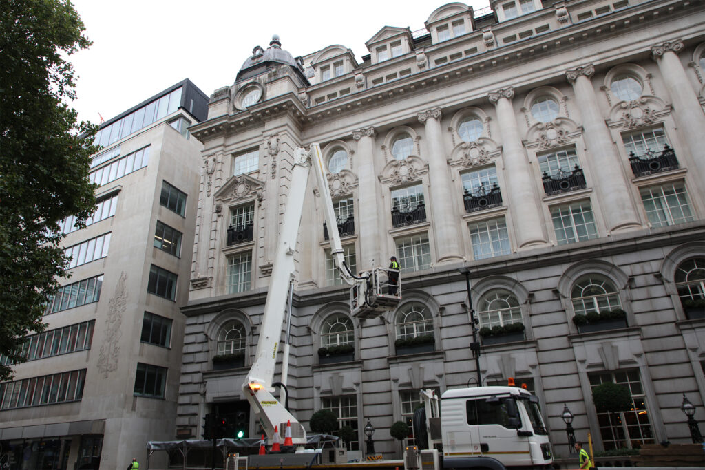 The ornate Portland stone façade of the Rosewood London hotel, Holborn, during a façade condition survey by Styles Façade Solutions (formerly Thomann-Hanry®). A surveyor in a cherry picker access platform, mounted on a white truck-mounted boom, is raised against the grand Edwardian Baroque elevation. The multi-storey façade features giant Corinthian columns, elaborate carved detailing, arched windows, wrought iron balconies and a copper-domed roofline. Traffic cones are visible at street level.