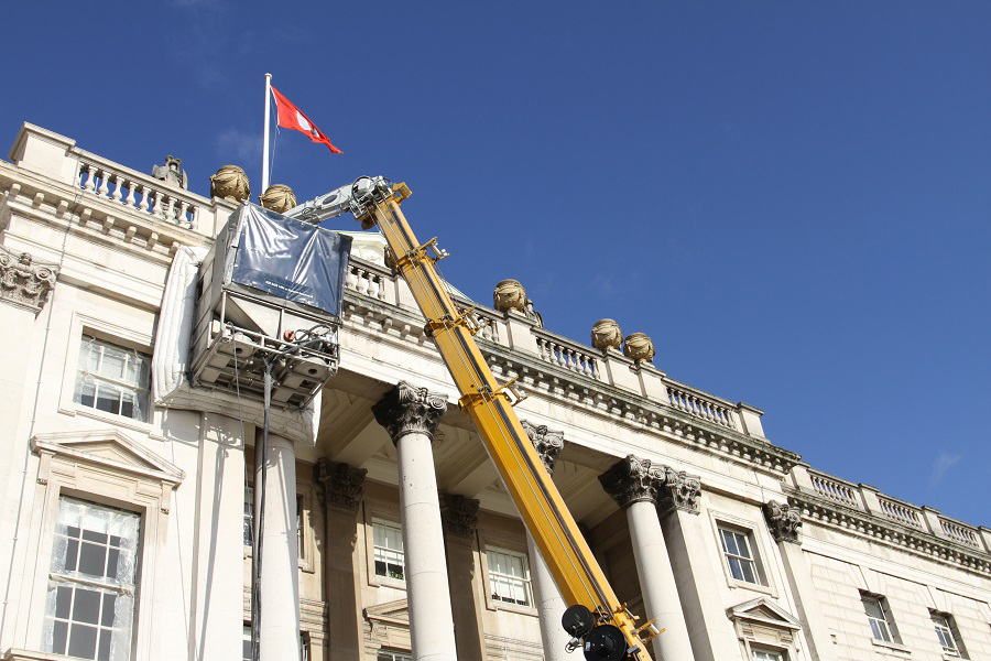 Classical Portland stone façade with fluted Corinthian columns, balustraded parapet, and decorative urns against a clear blue sky, with a red flag flying above. The crane with cleaning cabin attached is raised against the building. This picture captures the Façade Gommage cleaning process in progress, conducted by Styles Façade Solutions (formerly Thomann-Hanry®).