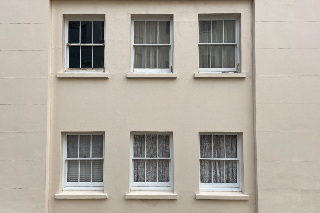 Six white-framed sash windows on a beige rendered façade prior to restoration, arranged in two rows of three. The upper-left window shows visible paint deterioration and exposed timber on its lower frame, while other windows display varying degrees of weathering. Net curtains and shutters are visible through several panes, with one window revealing a darker interior space beyond.