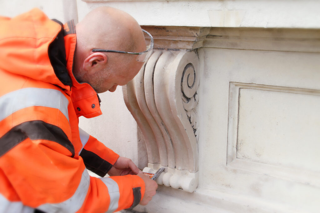 Styles Façade Solutions® carries out detailed stonemasonry on the Gibbs Building, King's College, Cambridge. A craftsman carefully carves a decorative scroll corbel by hand, demonstrating precise, skilled restoration work on the building's classical stone detailing without the use of scaffolding.