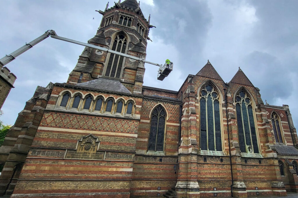Styles Façade Solutions® conducts a scaffold-free façade survey of the Grade I listed Rugby School Chapel. An operative on an elevated platform closely inspects the tower's decorative brickwork and stonework, assessing condition high above the Gothic Revival building's arched windows and pinnacles.