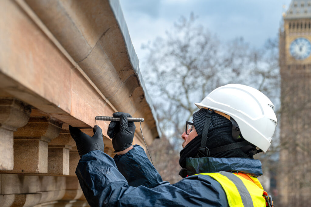 Close-up of a surveyor in a white hard hat, high-visibility vest and safety glasses closely inspecting carved stone cornice detailing on Surveyors House, London, home to RICS. Wearing protective gloves, he holds a metal probing tool against the weathered sandstone, checking for cracks and deterioration. The Elizabeth Tower, housing Big Ben, is visible in the blurred background, confirming the Westminster location of this façade survey.