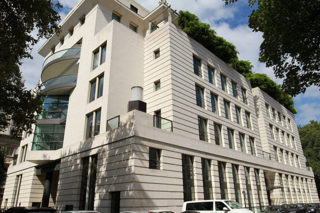 A striking contemporary corner building with pale cream stone cladding, featuring a distinctive curved glazed balcony, angular projecting bays, and a rooftop garden with lush greenery cascading over the parapet. Multiple rows of tall windows with dark frames punctuate the façade, and the stonework appears bright, clean, and uniformly toned throughout, reflecting the long-lasting protection provided by treatment with Aqua Fend®. Mature trees frame the scene under a partly cloudy blue sky, with parked cars visible at street level.
