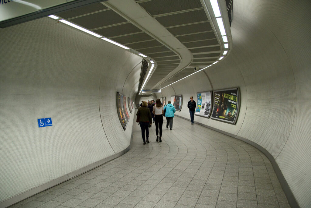 A curved, tiled underground walkway leading toward the Victoria line, its clean tiled floor treated with Aqua Fend® G. Pedestrians walk along the tunnel, passing illuminated film posters and advertisements lining the white-tiled walls, with a wheelchair access sign visible on the left. The floor's even, well-maintained appearance, free from staining or discolouration despite heavy footfall, demonstrates the lasting protective benefits of treatment against urban grime and wear in this busy transport environment.