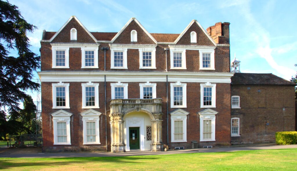 Front view of Boston Manor House, London, a red-brick Jacobean building with three prominent gables and white-framed sash windows arranged symmetrically across three storeys. An ornate stone-columned porch frames the green front door beneath "BOSTON MANOR HOUSE" signage. A tall brick chimney stack and small lantern cupola rise above the roofline, with a cedar tree and open lawn in the foreground beneath a blue sky. Boston Manor House: Photo Credit P.G.Champion CC BY-SA 2.0