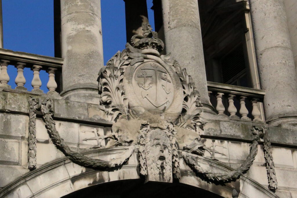 Ornate carved stone crest on the Grade I listed Somerset House, London, prior to cleaning with the Façade Gommage process by Styles Façade Solutions®. The crowned shield bears two crossed anchors, surrounded by acanthus leaf scrollwork. Below, a bearded human face forms part of the keystone, flanked by carved swags looping across the archway to smaller masks on either side. Weathered columns and a balustraded balcony rise behind. The stonework shows moderate soiling, with darker staining and deposits built up within the carved recesses and undersides of the ornamentation.