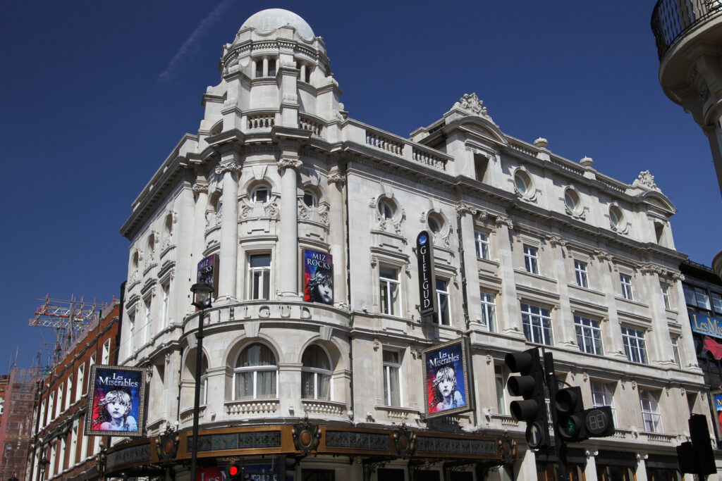 The Gielgud Theatre, London, an ornate white stone building with a domed corner turret, decorative columns, and rounded oval windows. Large "Les Misérables" and "Miz Rocks" posters adorn the façade beneath the "GIELGUD" signage, with a hanging "Gielgud Theatre" sign extending from the corner. Traffic lights and street lamps stand in front, set against a bright blue sky at this busy West End corner.