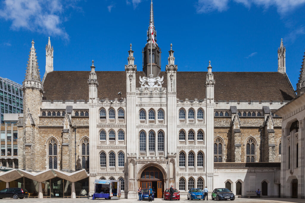 Front-facing view of Guildhall, London, showcasing its Gothic façade with tall arched windows, ornate stone pinnacles, and a central tower topped with a flagpole flying the St George's cross. The heraldic griffin crest and "DOMINE DIRIGE NOS" inscription sit above the main arched entrance. Cars and pedestrians occupy the paved courtyard in front, set against a bright blue sky. Guildhall: Photo Credit Diego Delso CC BY-SA 4.0