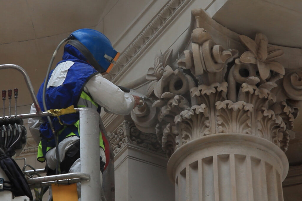 Styles Façade Solutions® performing delicate Façade Gommage cleaning on an intricately carved Corinthian capital at the Grade I listed Bank of England. A fully protected operative uses the heritage grade Façade Gommage cleaning process to carefully remove soiling from the acanthus leaf detailing, preserving the stonework's fragile carved heritage.
