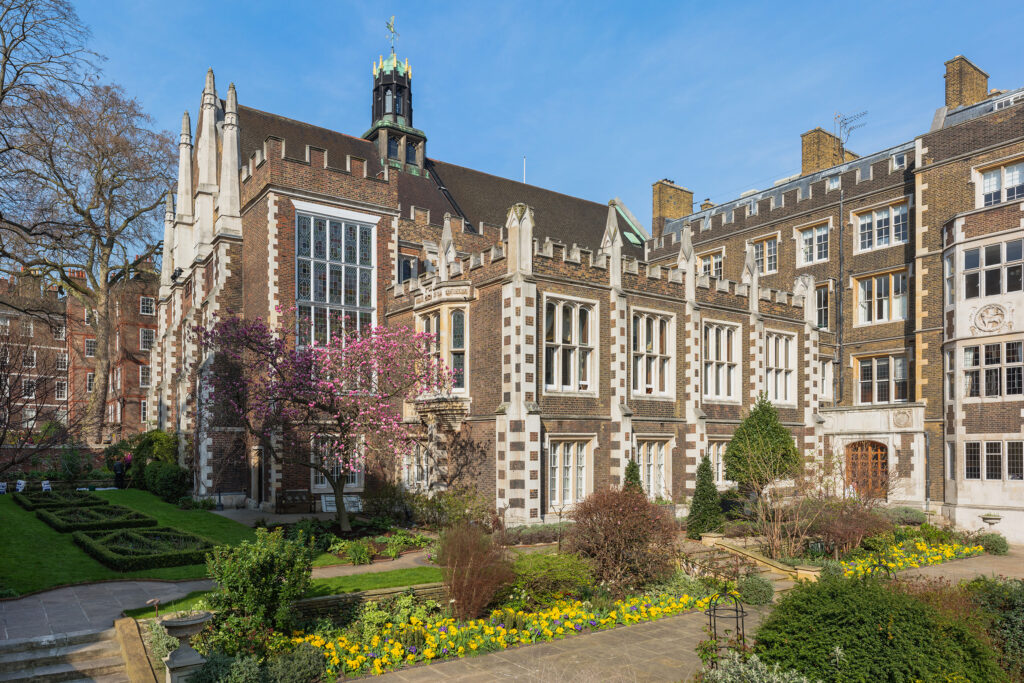 Middle Temple Hall and surrounding buildings, part of the Inns of Court, London, featuring Tudor-style brick and stone architecture with mullioned windows, crenellated parapets, and a distinctive black lantern tower. A flowering pink magnolia tree and manicured garden with box hedges, yellow spring flowers, and lawns sit in front of the historic buildings beneath a clear blue sky. Middle Temple Inns of Court: Photo Credit Diliff CC BY-SA 3.0