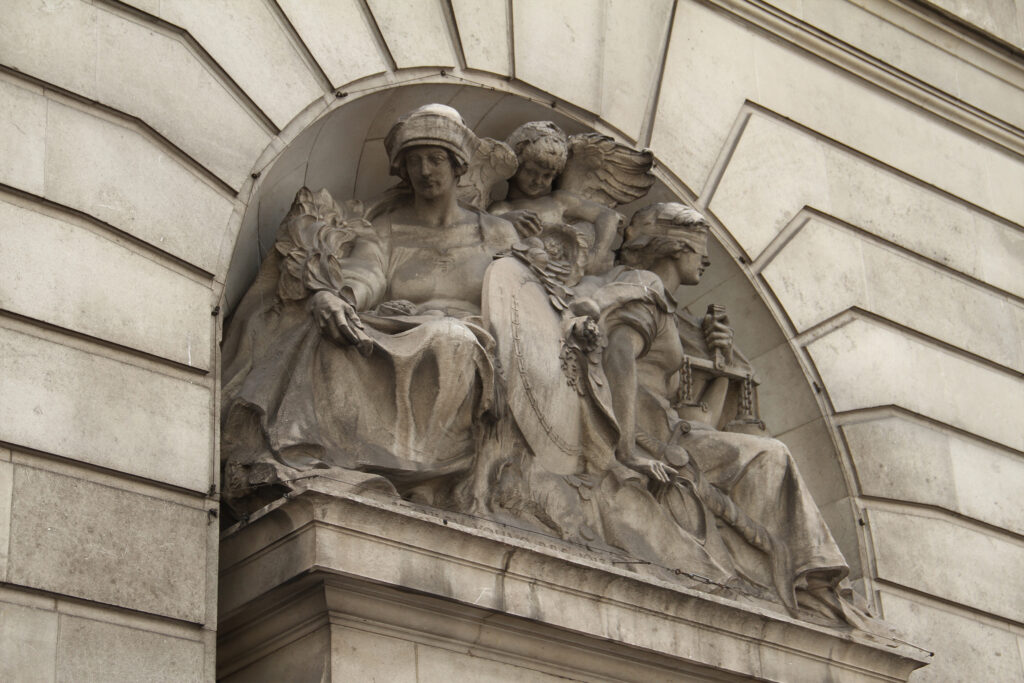Weathered Portland stone sculptural relief depicting allegorical figures within an arched niche, showing visible grime and dark staining across the carved surfaces. A helmeted figure, winged cherub, and draped woman holding scales sit amid ornate stonework, their once-crisp details softened by pollution build-up. The surrounding pale stone façade contrasts against the darker, soiled carving, illustrating the effects of urban weathering.