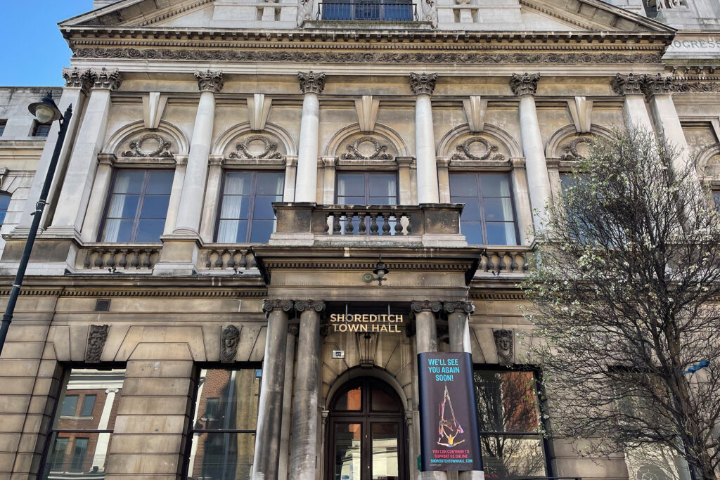 Front façade of Shoreditch Town Hall, London, before cleaning, showing weathered and darkened Portland stone with heavy grime staining across the columns, arched windows, and carved ornamental details. The building's grand Corinthian columns and decorative balustrade appear discoloured, with "SHOREDITCH TOWN HALL" signage above the entrance. A blooming tree stands beside the building, contrasting fresh spring growth against the soiled stonework beneath a clear blue sky.