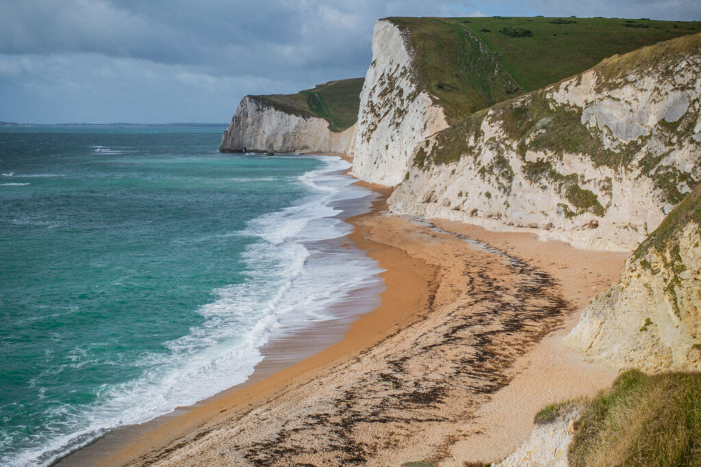 Dramatic view of the Jurassic Coast, Dorset, showing towering white limestone cliffs curving along a golden sandy beach beside turquoise sea waters. Waves break gently along the shoreline, with seaweed lines marking the tide's edge on the sand. Grassy clifftops stretch into the distance under a moody, cloud-filled sky, showcasing the rugged natural beauty of this stretch of English coastline.