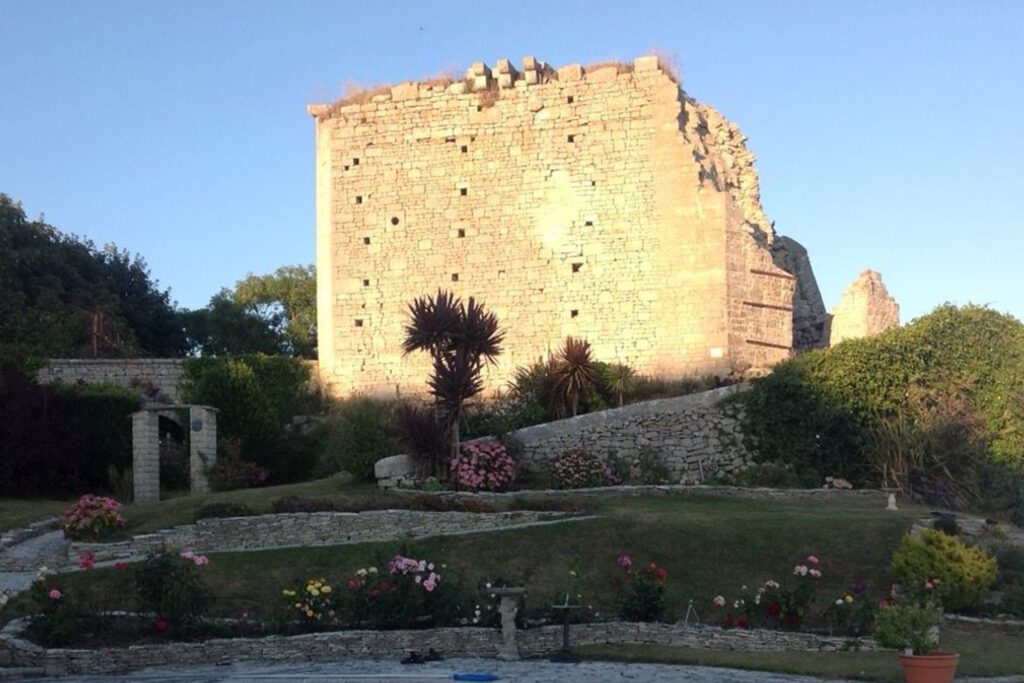 Rufus Castle ruins at Church Ope Cove, Portland, Dorset, its weathered golden stone walls rising dramatically against a clear blue sky. The crumbling square tower shows visible cracks and missing sections, with small square window openings dotting the ancient stonework. A landscaped garden with terraced stone walls, colourful flowerbeds, and a palm tree sits below, blending historic ruins with cultivated greenery in the evening light. Photo Credit: Artefax CC BY-SA 4.0