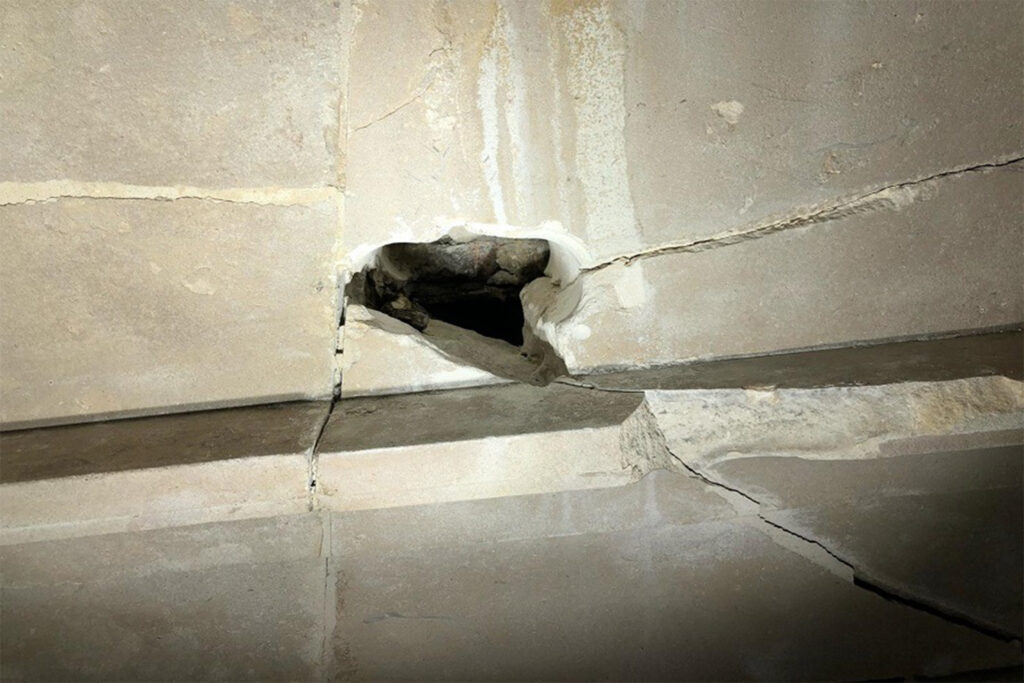 Close-up of "Regent Street Disease" affecting a stone building façade, showing a fractured cavity where corroded metal cramps have expanded and split the Portland stone. Rust-coloured staining streaks down from the damaged joint, while hairline cracks run along the stonework above and below. The exposed void reveals crumbling stone and corrosion product, illustrating the structural deterioration this condition causes over time.