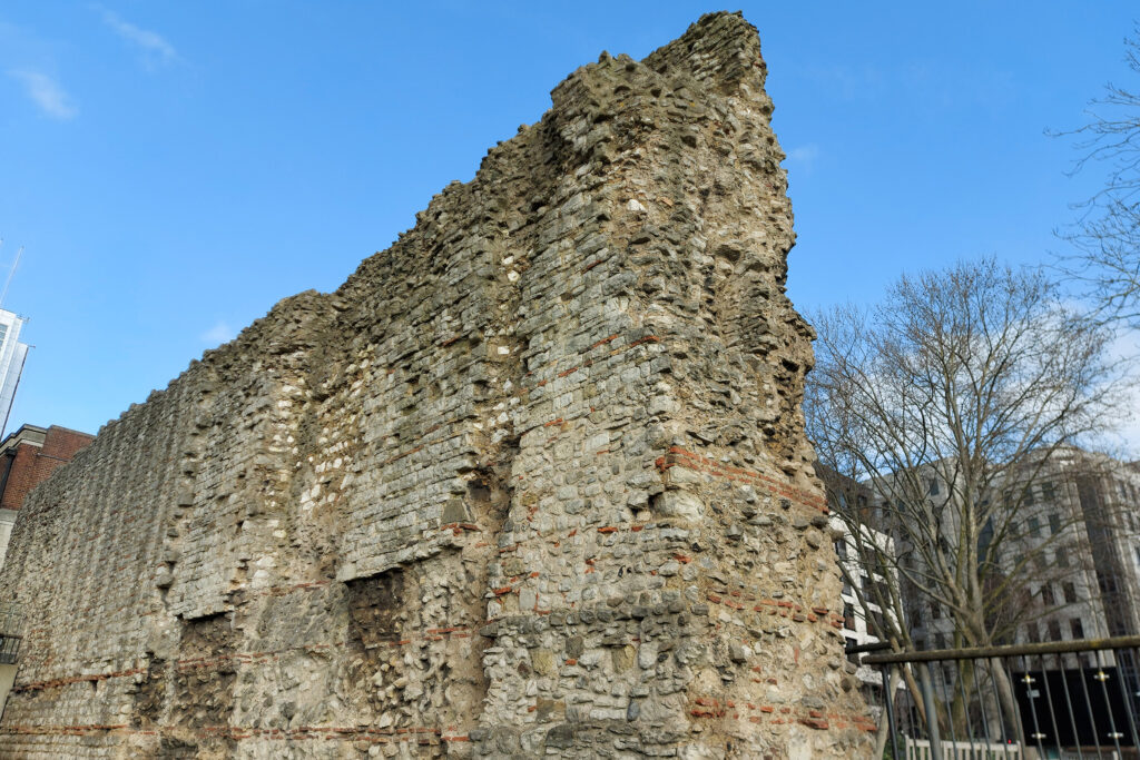 A surviving section of London's ancient Roman Wall, standing tall against a clear blue sky. The weathered stone structure shows layers of ragstone rubble interspersed with distinctive red brick courses, its jagged, eroded top testament to centuries of exposure. Bare trees and modern glass and brick buildings frame the historic ruin, highlighting the contrast between ancient and contemporary London. Ruins of the Roman Wall: Photo Credit Jamzze CC BY-SA 4.0