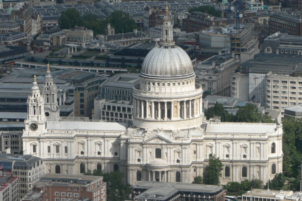 Aerial view of St Paul's Cathedral, London, showcasing its iconic domed roofline with a golden-tipped lantern and cross rising above the columned drum. Twin baroque bell towers stand at the west front, one displaying a clock face. The white Portland stone façade contrasts against surrounding London rooftops, glass office buildings, and scattered green trees, capturing the cathedral's dominant presence within the modern cityscape. St Paul’s Cathedral: Photo Credit Mark Fosh CC BY 2.0