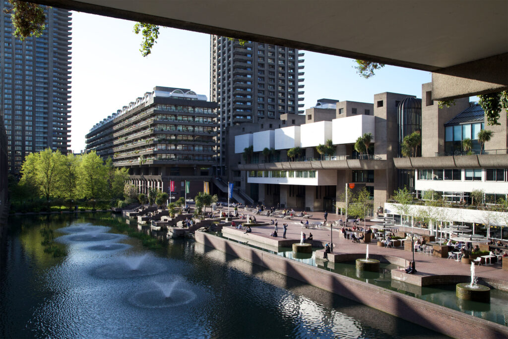 The Barbican Centre, London, showcasing its distinctive Brutalist architecture with raw concrete buildings and towering residential blocks rising in the background. A tranquil lakeside terrace features spouting fountains, potted palm trees, and a bustling waterside café where visitors relax at outdoor tables. Reflections shimmer across the water beneath a bright sky, capturing the striking contrast between concrete structures and lush greenery. The Barbican Centre: Photo Credit Tom Parnell CC BY-SA 2.0